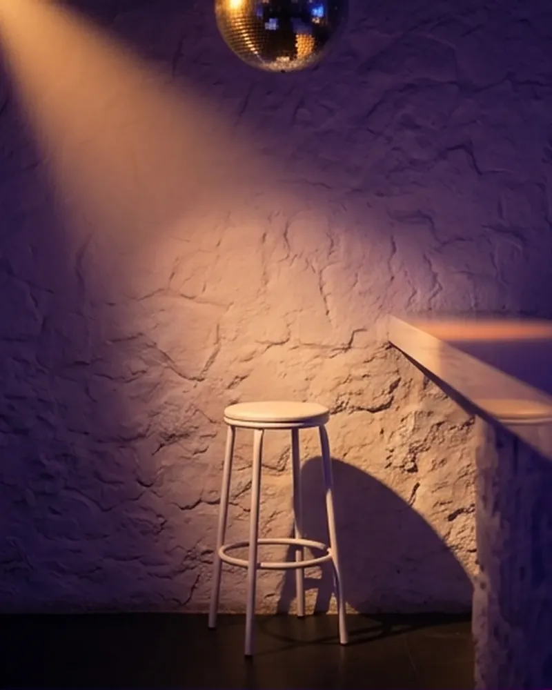 Taburete blanco junto a una pared de estuco, bajo un haz de luz cálida y la bola de espejos.